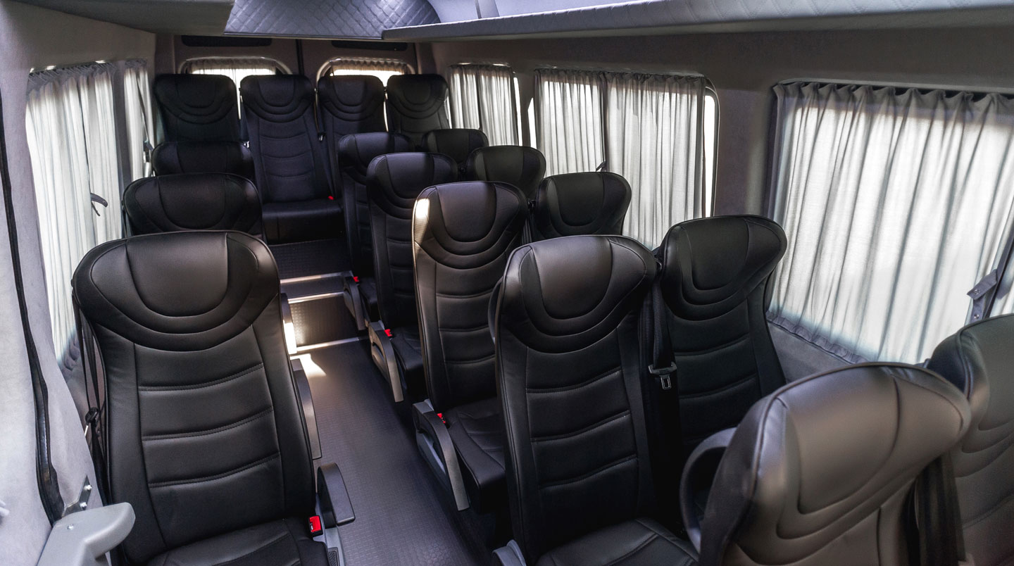 Interior of modern minibus showing three rows of upholstered passenger seats, overhead luggage storage, and large windows with natural daylight